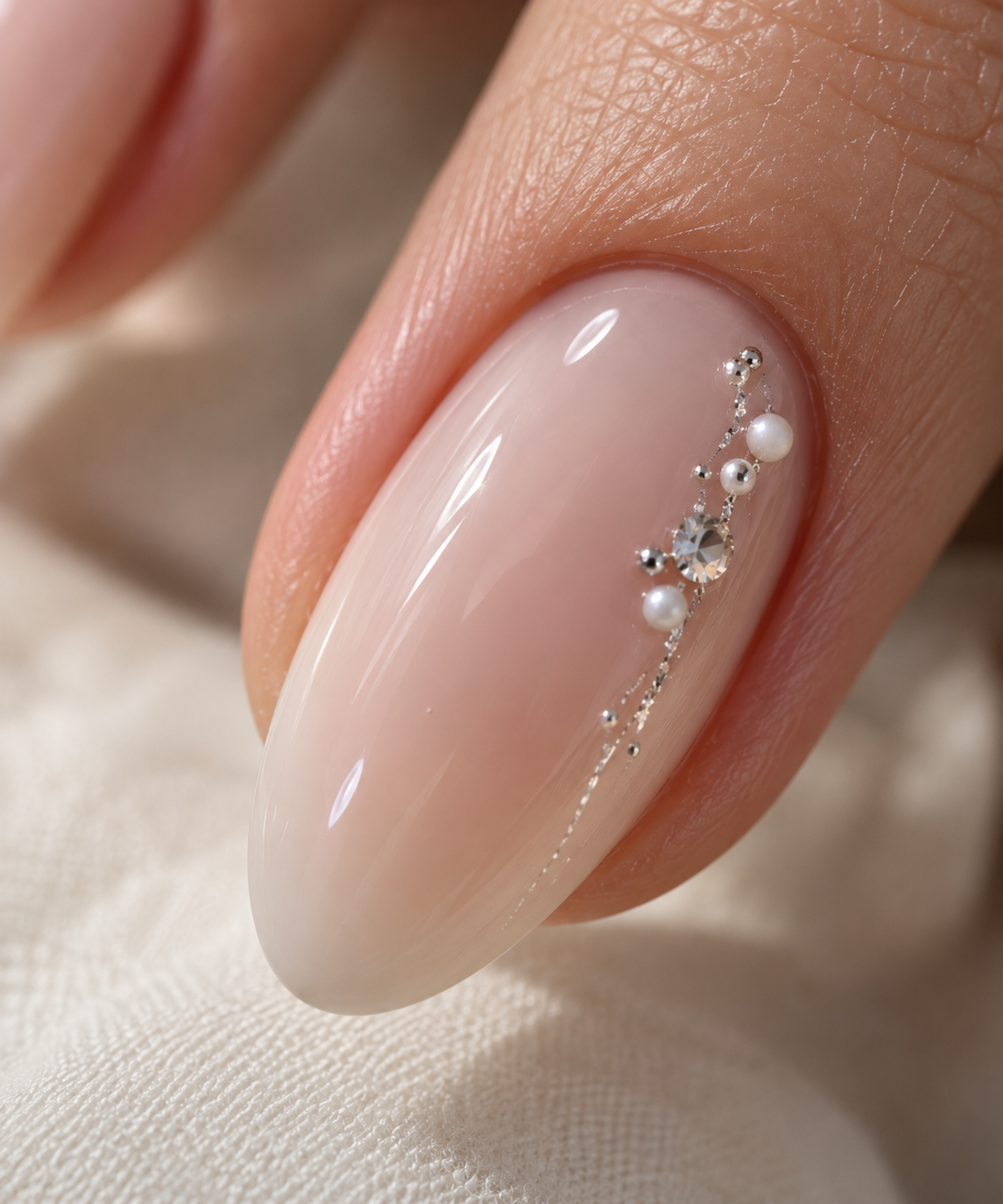 Gros plan sur un ongle amande nude pêche orné de perles nacrées, d’un cristal et d’une fine chaîne argentée, création Joaillerie de l’ongle pour la mariée